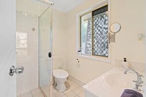 A bathroom with a glass shower enclosure, a toilet, a sink with a mirror, and a window with a decorative screen. - Riverview Modern Haven | Entire 4BR Home (Riverview)