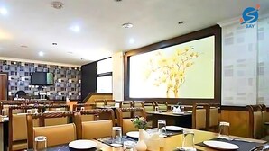 A dining area with a long table set for a meal, surrounded by cushioned chairs. A projector screen is mounted on the wall displaying a floral image. - Say Moments (Kolkata)