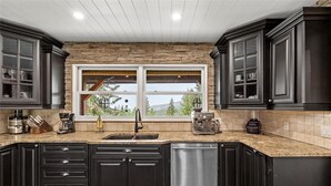A modern kitchen with dark cabinetry, a stone accent wall, a large window, and a stainless steel dishwasher.