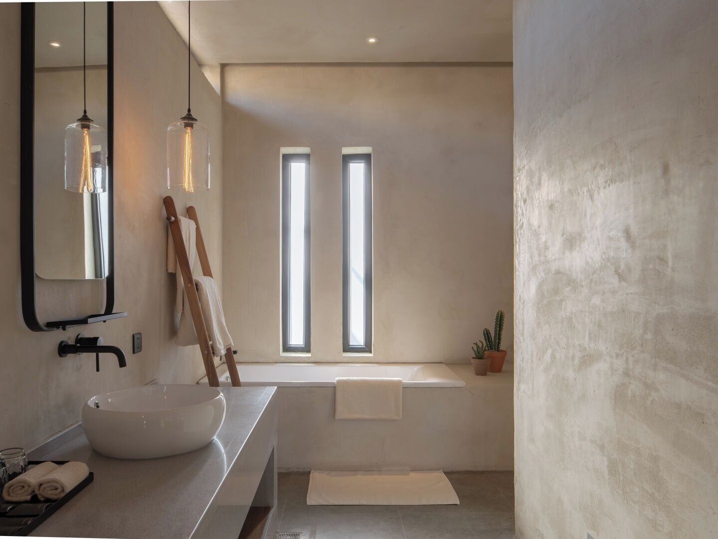 A modern bathroom with a large sink, a mirror, and a bathtub. There are two windows providing natural light, and a wooden ladder leaning against the wall.