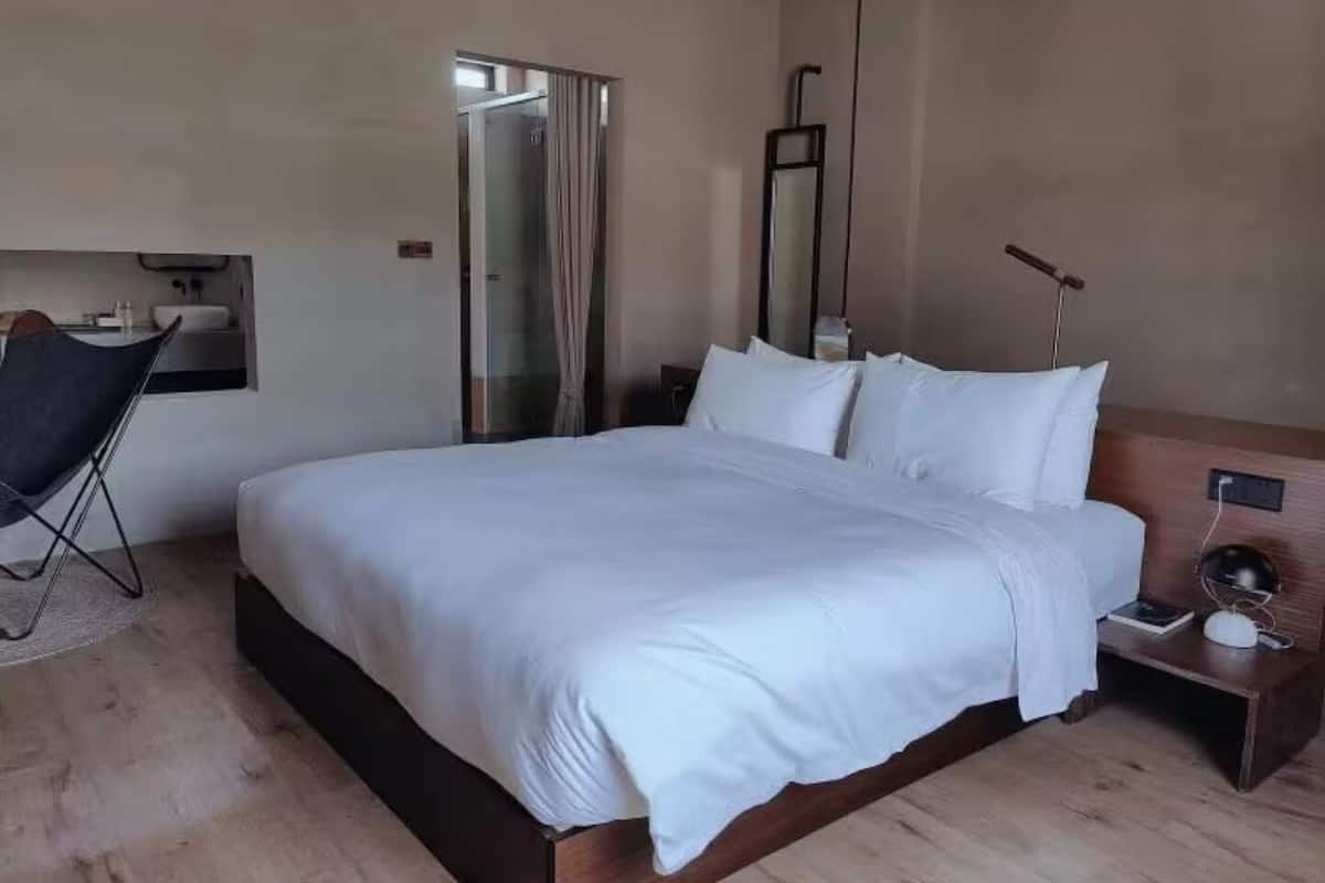 Une chambre d’hôtel moderne avec un grand lit, un petit bureau, une chaise et une salle de bain visible à travers une porte en verre.