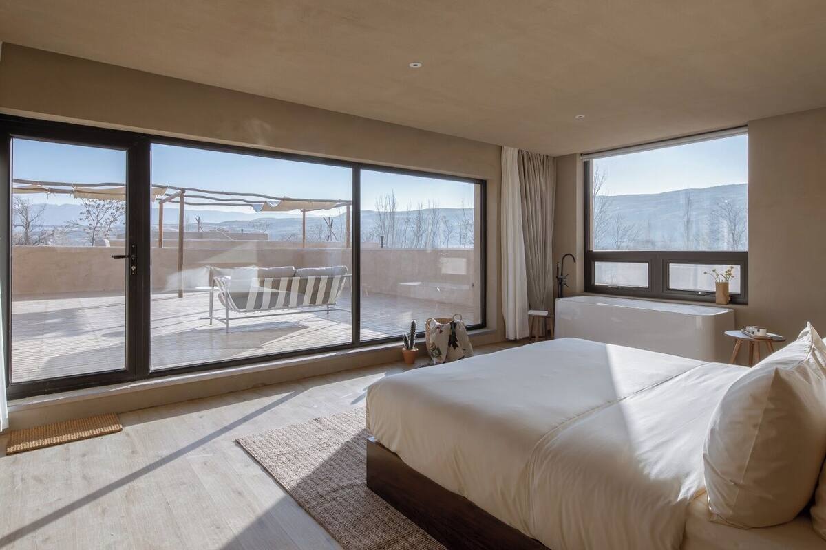 Une chambre moderne avec un grand lit, une vue sur un patio aménagé avec du mobilier d’extérieur et une grande fenêtre.