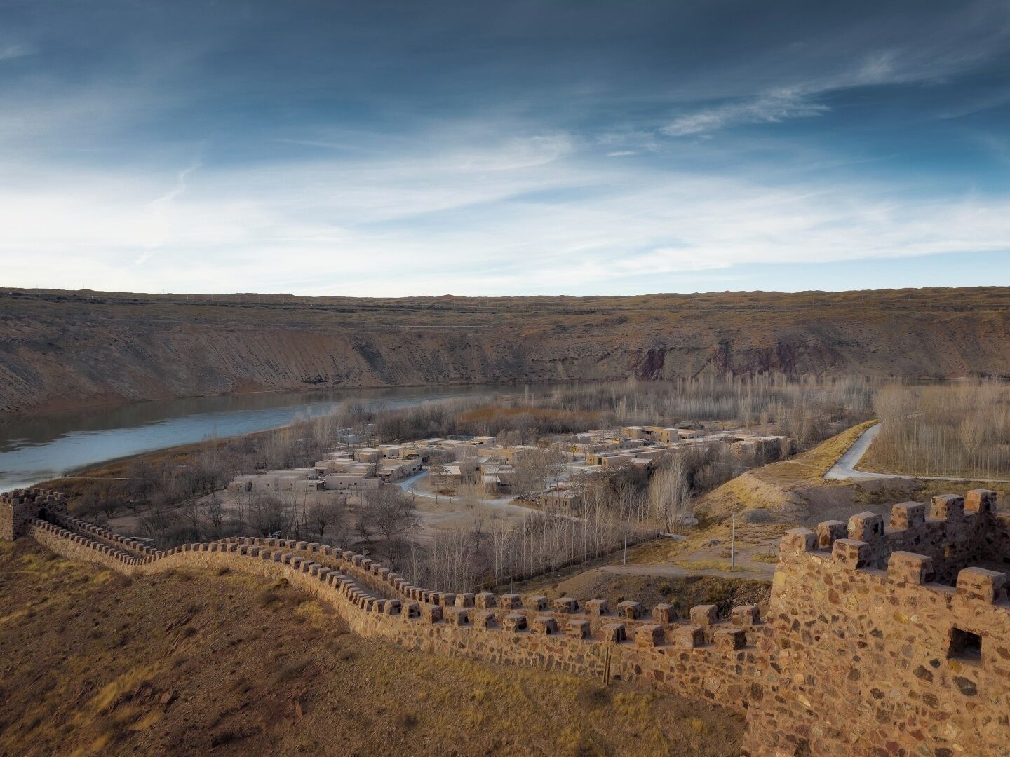 A historical site with a stone wall and ruins, surrounded by a river and sparse vegetation.