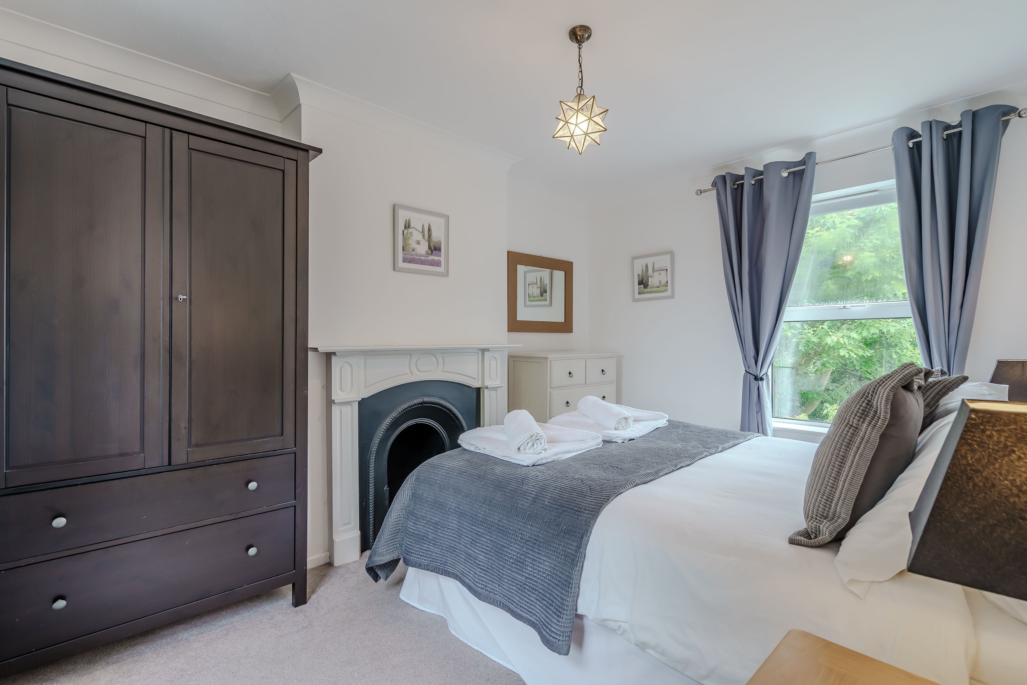 A bedroom with a large wardrobe, a fireplace, a bed with a grey blanket, and a window with curtains.