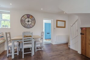 A dining area with a wooden table and chairs, a large wall clock, and a blue door in the background. - The Net House | Winterton Cottages (Winterton-on-Sea)