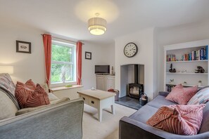 A living room with a sofa, a table, a fireplace, a bookshelf, and a clock on the wall. - The Net House | Winterton Cottages (Winterton-on-Sea)