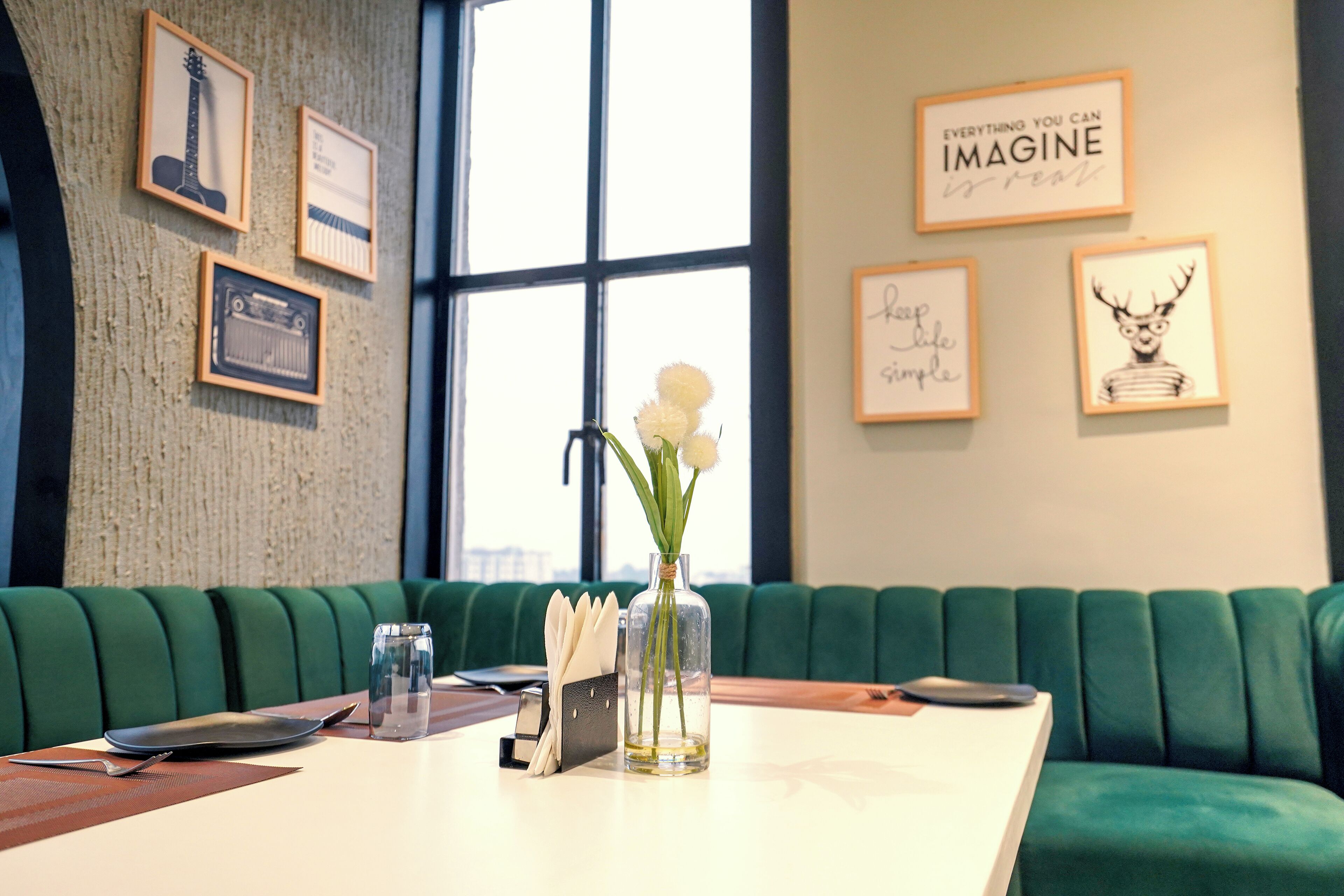 Un comedor con una mesa dispuesta para dos personas, un florero con flores, cuadros en la pared y un banco tapizado en verde.