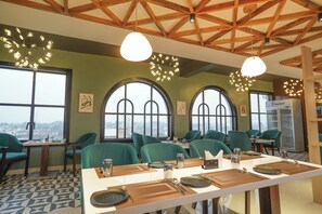 Ein Restaurant mit hohen Holzbalkendecken, großen Bogenfenstern und smaragdgrünen Stühlen. Die Tische sind mit Tellern, Besteck und Gläsern gedeckt.
