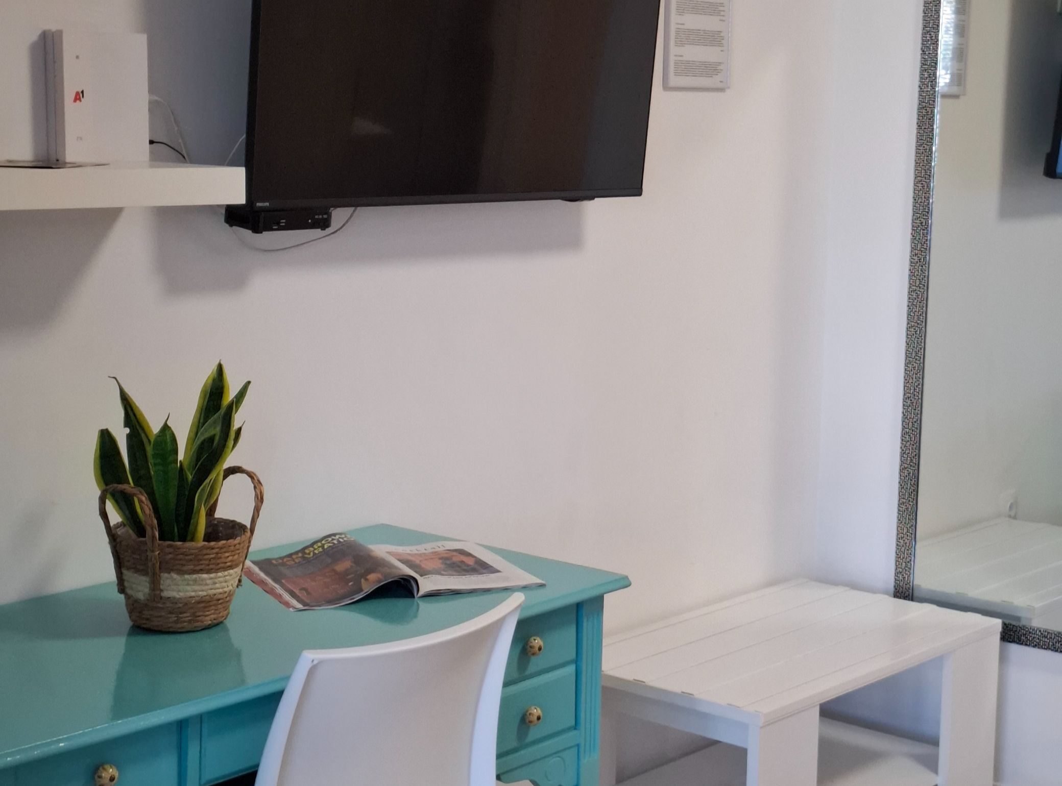 Una habitación con un escritorio verde azulado, una silla blanca, un televisor montado en la pared y una planta pequeña en una cesta.