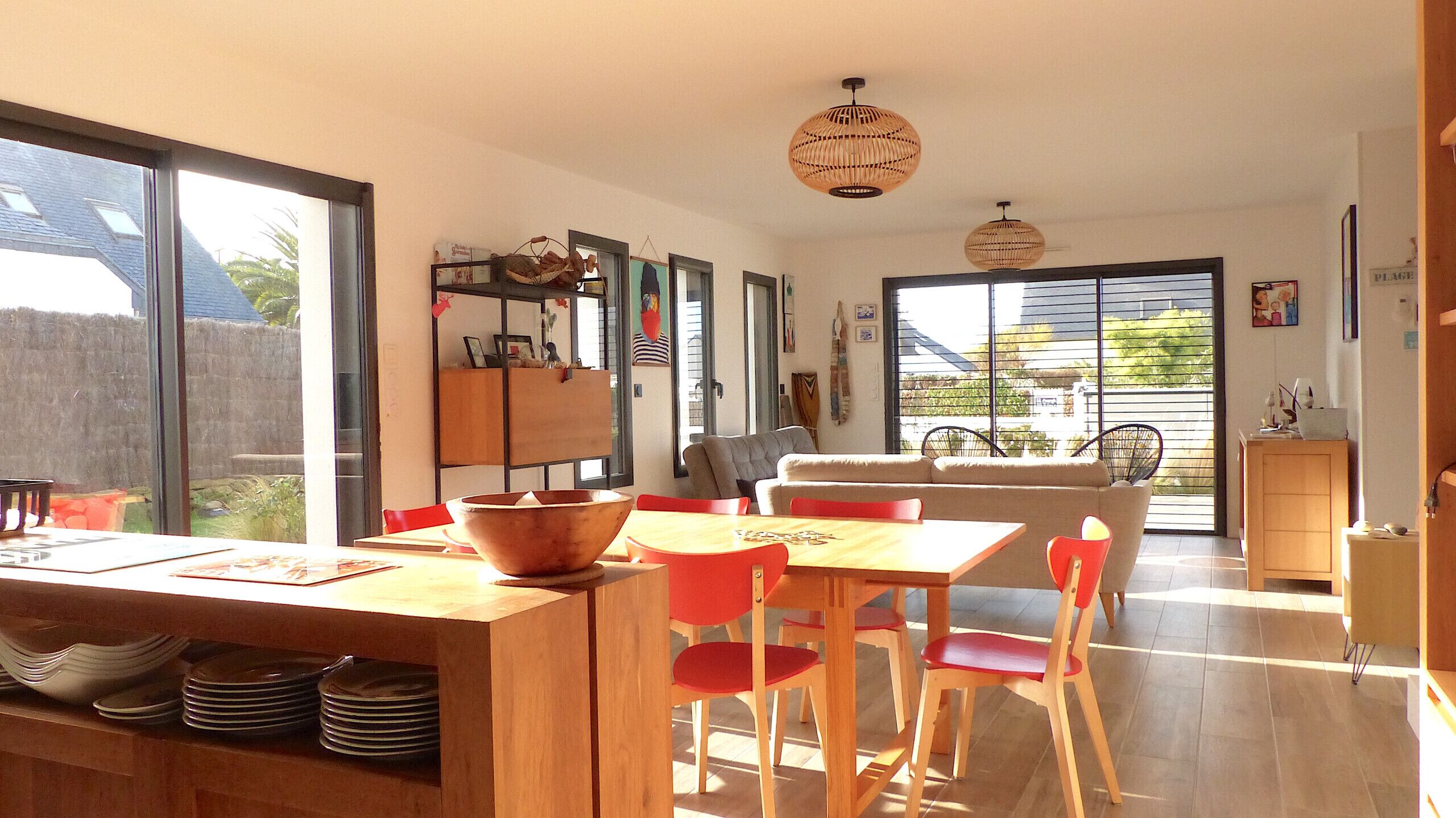 Un comedor luminoso con una mesa de madera y sillas rojas, una cocina con un ventanal grande, y una sala de estar con sofá y sillones.