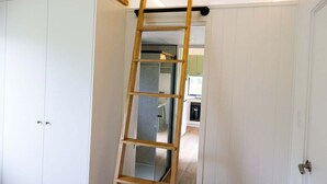 Une pièce avec une échelle en bois appuyée contre une porte blanche, une étagère en bois au-dessus de la porte, et on aperçoit une cuisine au fond.