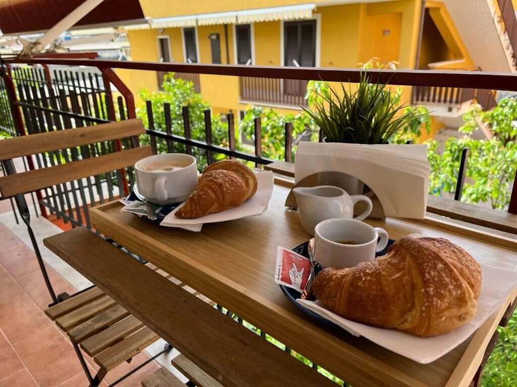 Una mesa de madera con croissants, tazas de café y una planta pequeña en un balcón con vistas a un edificio amarillo.