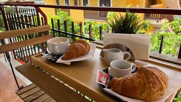 Una mesa de madera con croissants, tazas de café y una planta pequeña en un balcón con vistas a un edificio amarillo.