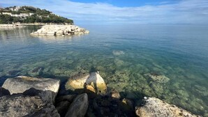 Paesaggio costiero con acque limpide, formazioni rocciose e una collina lontana con edifici.