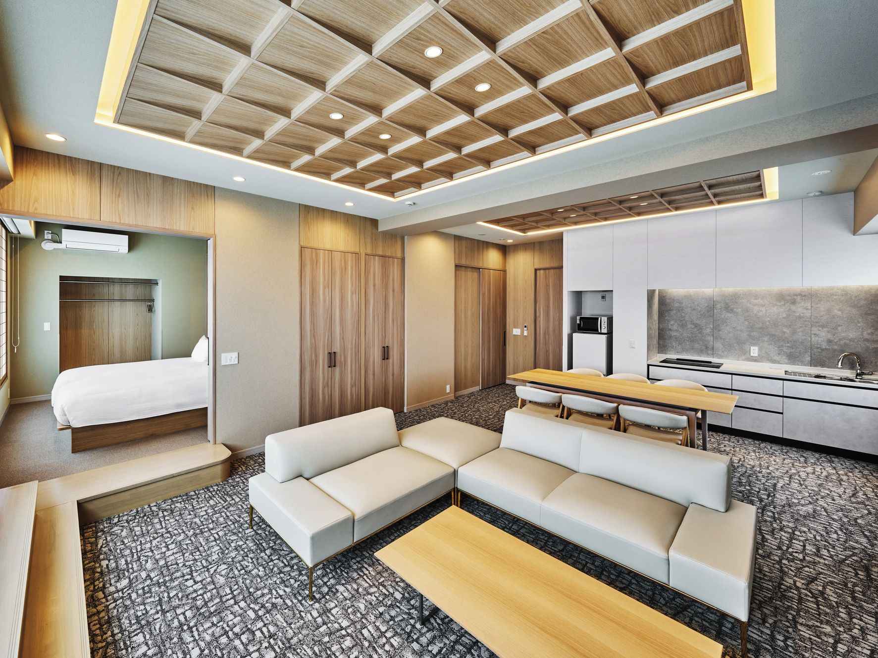 A modern hotel room with a bed, sofa, dining table, kitchenette, and a large window.