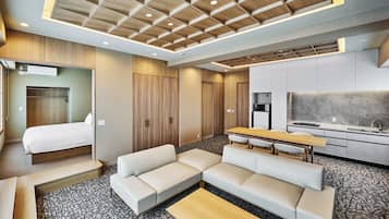 A modern hotel room with a bed, sofa, dining table, kitchenette, and a large window.