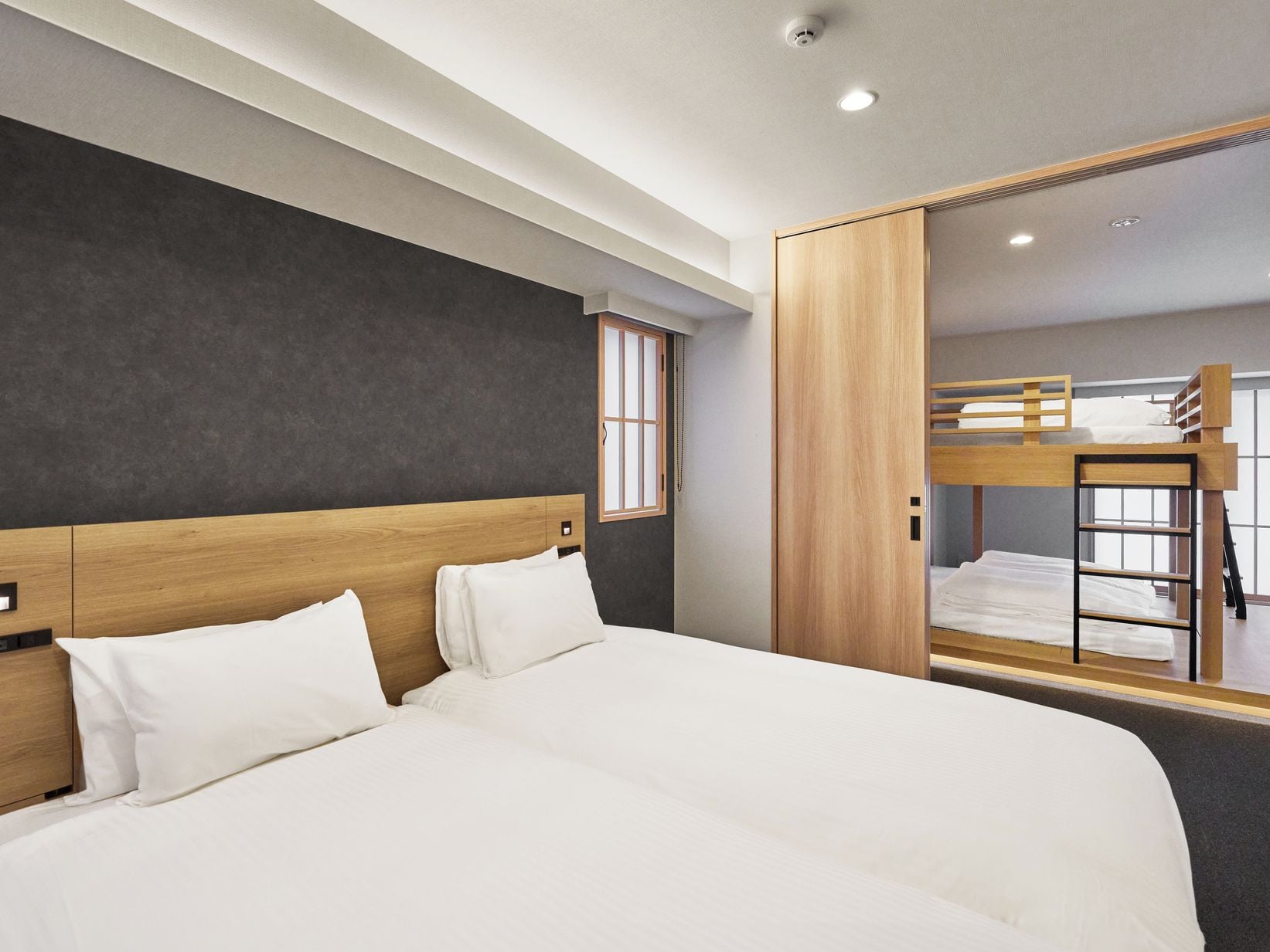 A modern hotel room with a large bed, a wooden headboard, a wardrobe, and a bunk bed in the background.