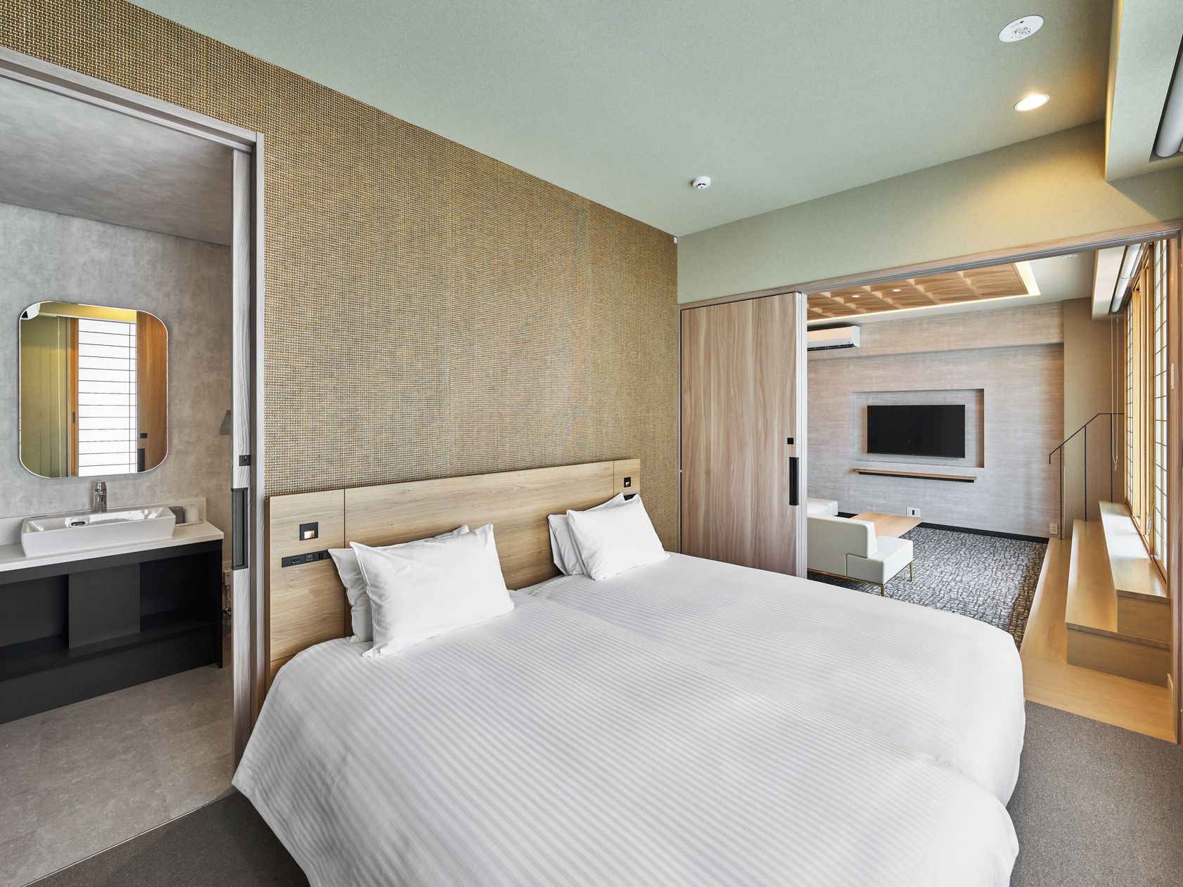 Ein modernes Hotelzimmer mit einem großen Bett, einem Badezimmer inklusive Waschbecken und Spiegel, einem an der Wand befestigten Fernseher und einem Holzkopfteil.