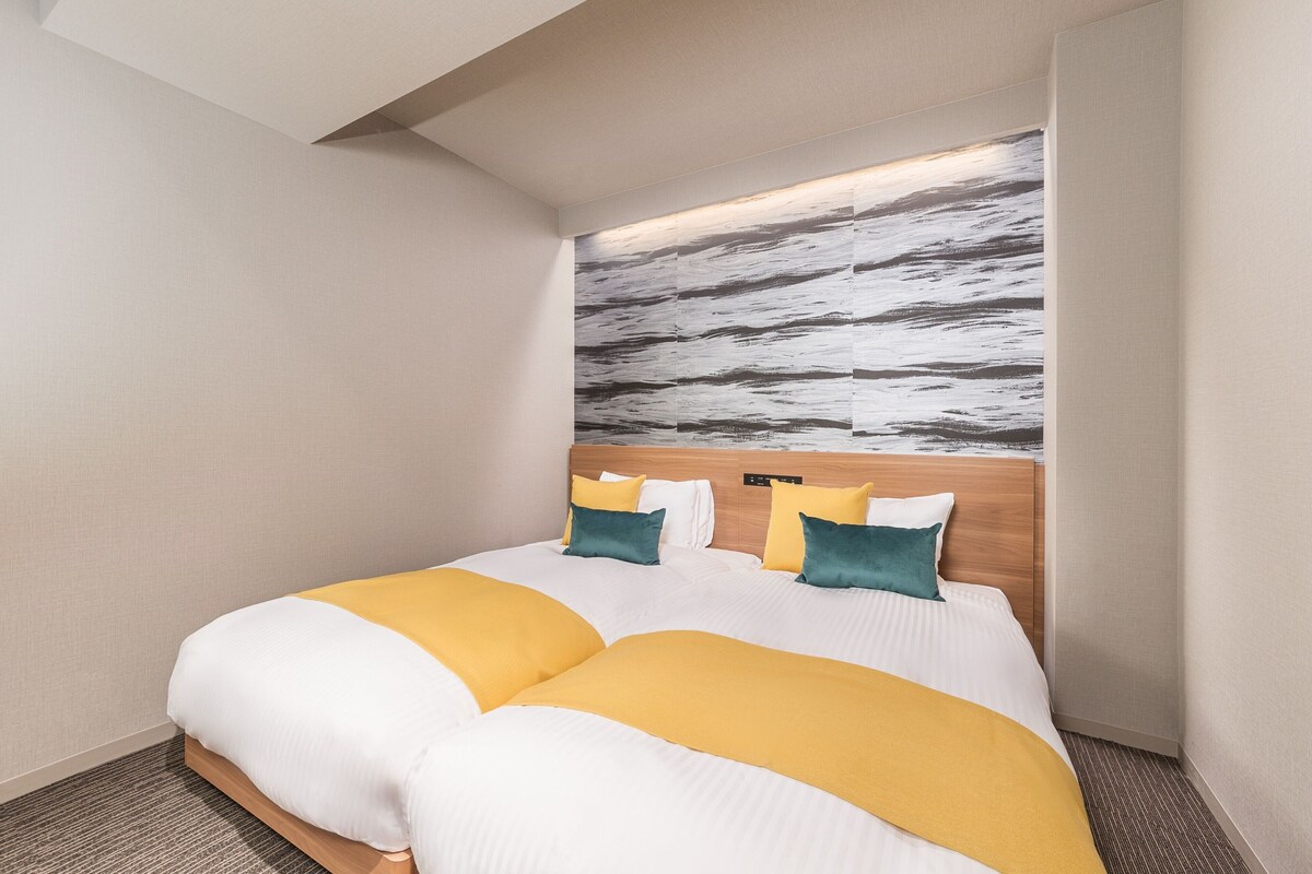 A modern hotel room with a large bed, a headboard with a control panel, and a textured wall art above the bed.