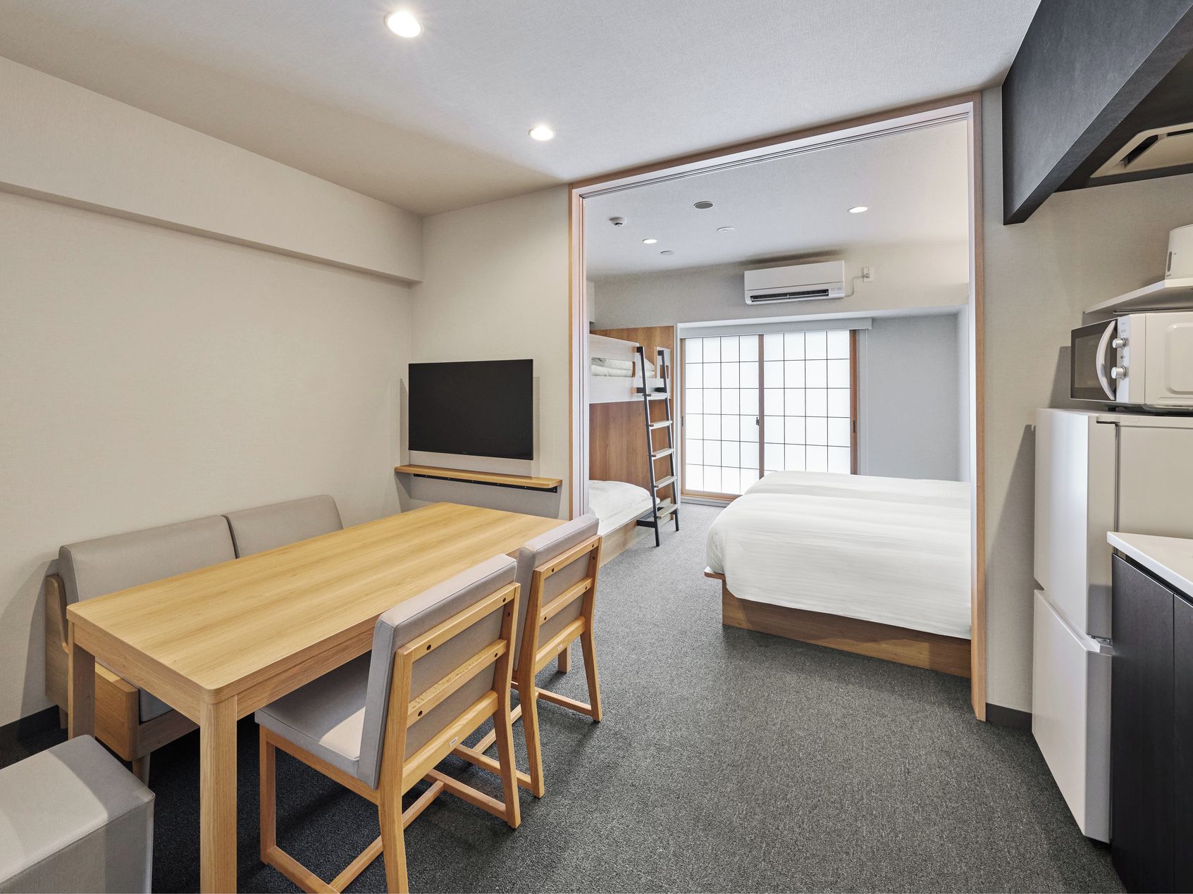 A compact hotel room with a wooden dining table, a bed, a kitchenette, and a television.