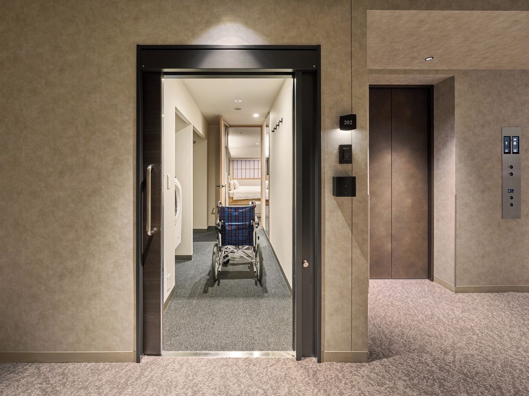 An elevator with a control panel, a wheelchair, and a corridor leading to a room.