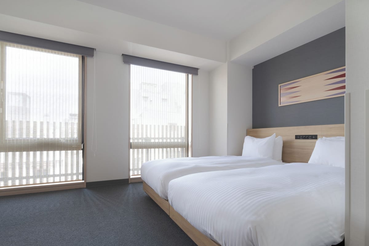A hotel room with a large bed, two windows with blinds, a headboard with a control panel, and a painting on the wall.