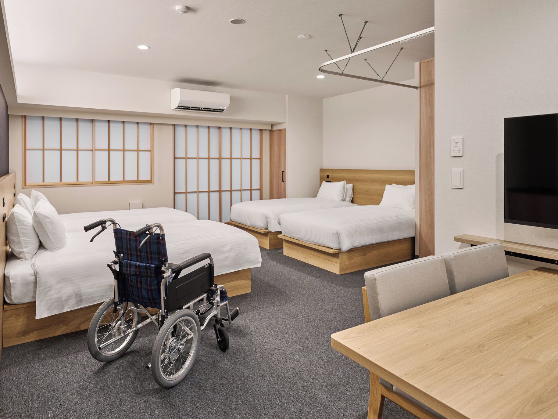 A hotel room with two beds, a wheelchair, a TV, and a dining table.