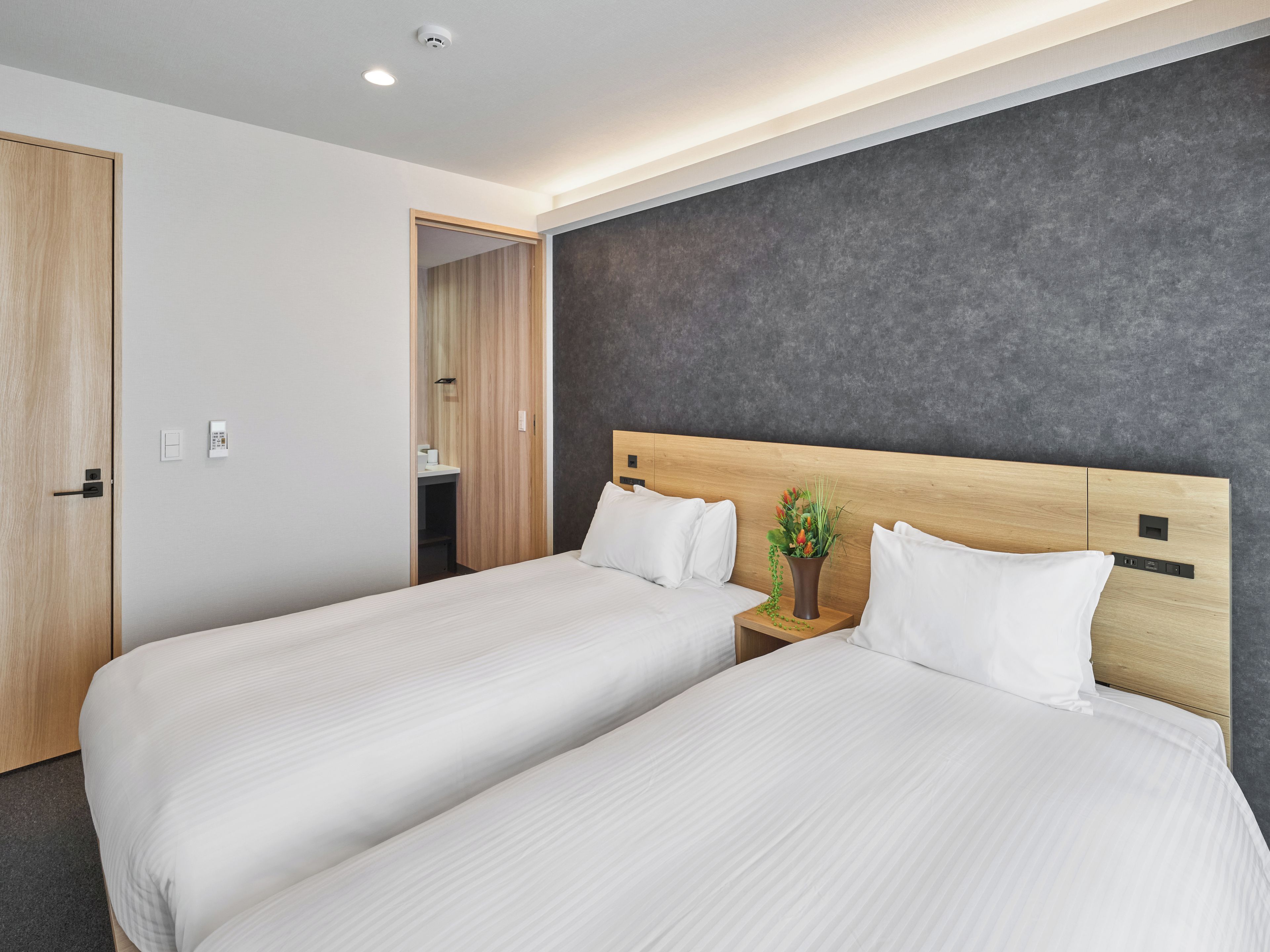 Una habitación de hotel moderna con dos camas, un cabecero de madera, una pared de acento gris y una mesita de noche con una planta.