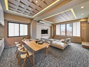A modern hotel room with a wooden ceiling, a dining area with a table and chairs, a sofa, and large windows.