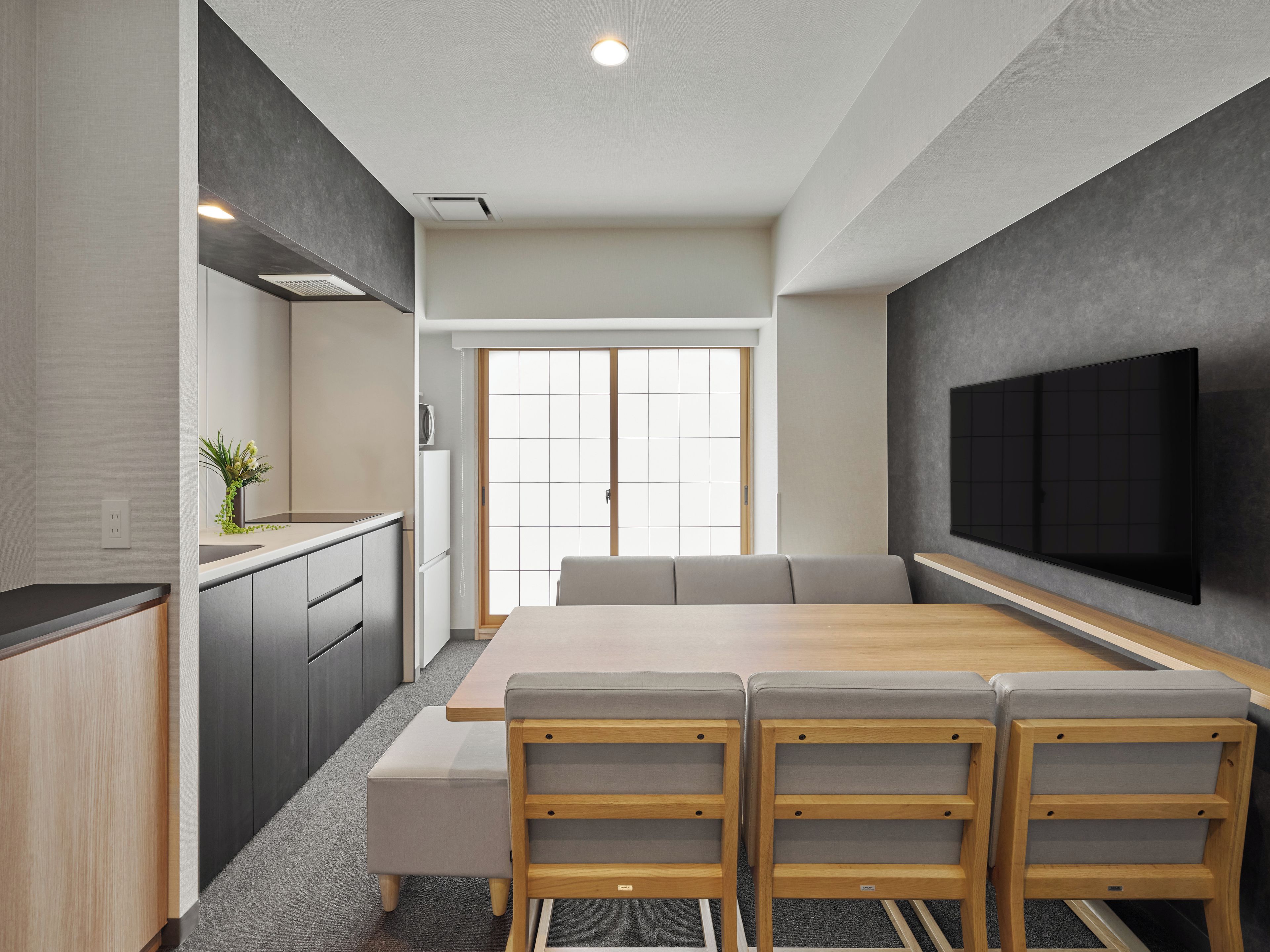 A modern kitchen with a dining area, a flat-screen TV mounted on the wall, and a sliding door leading to a bright room.