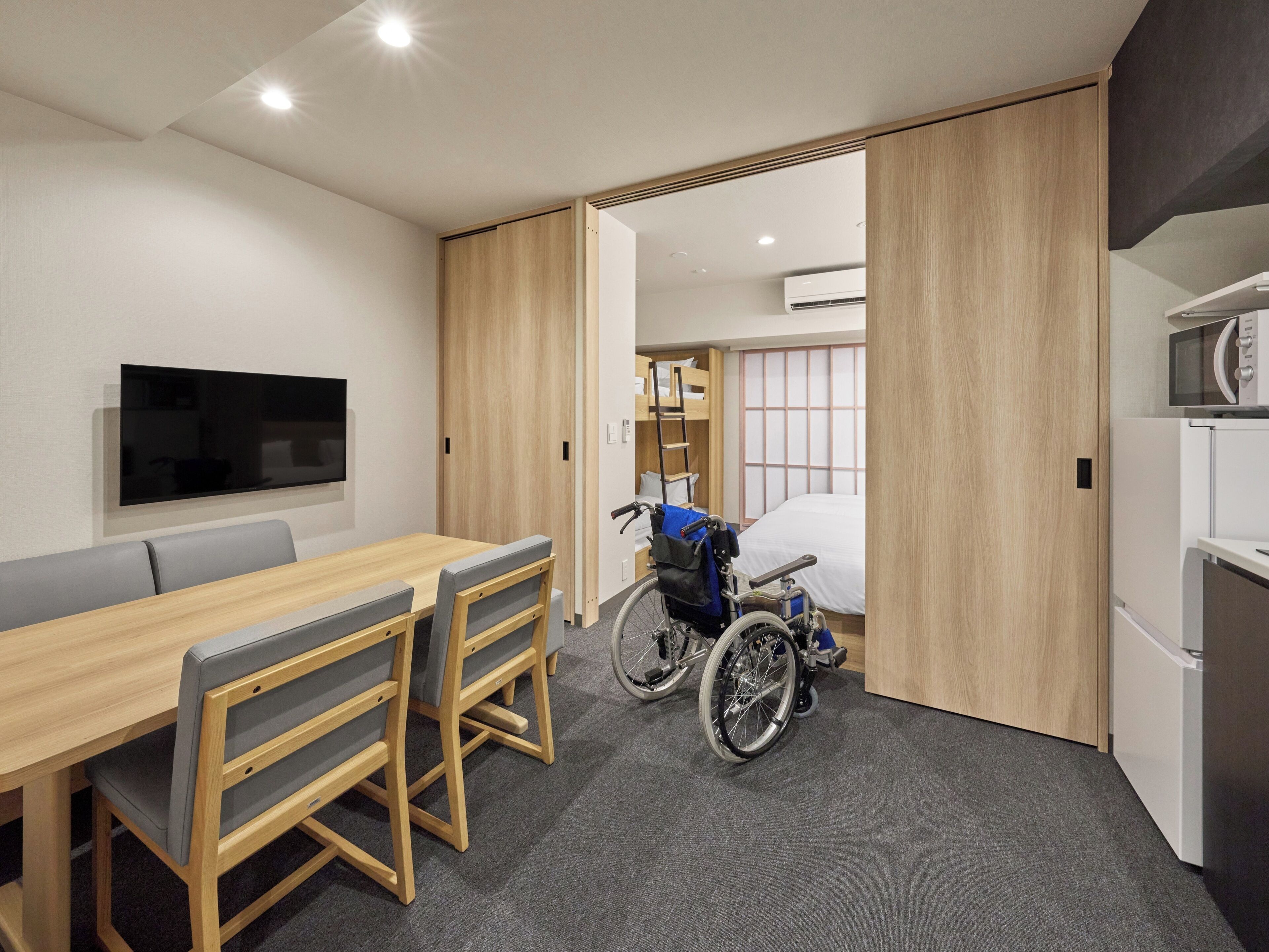 A modern room with a wooden dining table, chairs, a wheelchair, and a bed with a bunk.