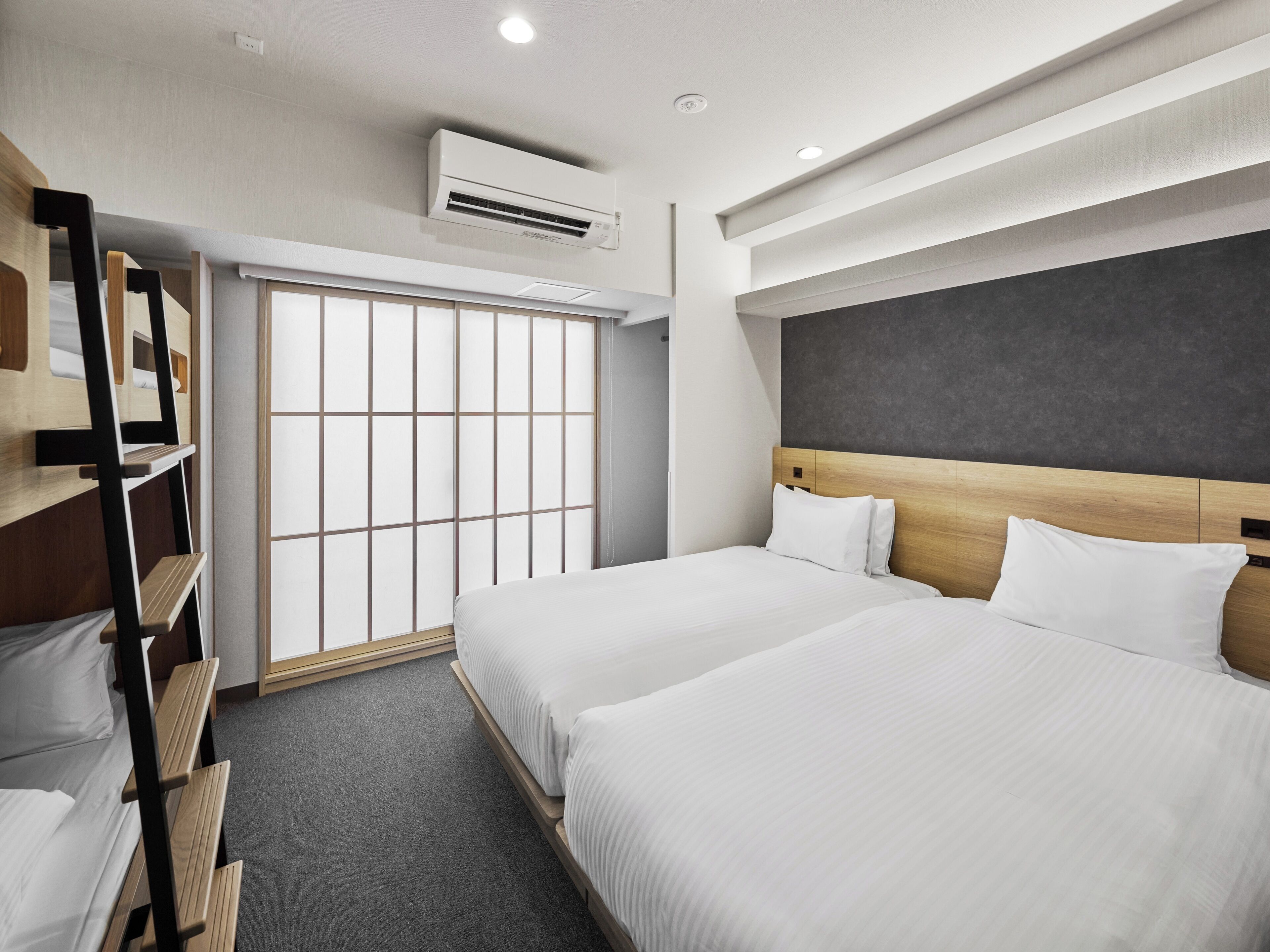 Habitación de hotel con dos camas, un armario, una ventana con persianas, un equipo de aire acondicionado y una lámpara en el techo.