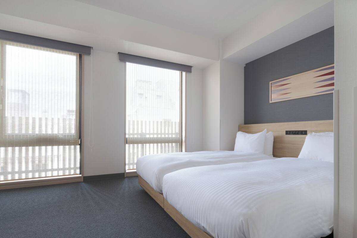 A hotel room with a large bed, two windows with blinds, a headboard with a control panel, and a painting on the wall.