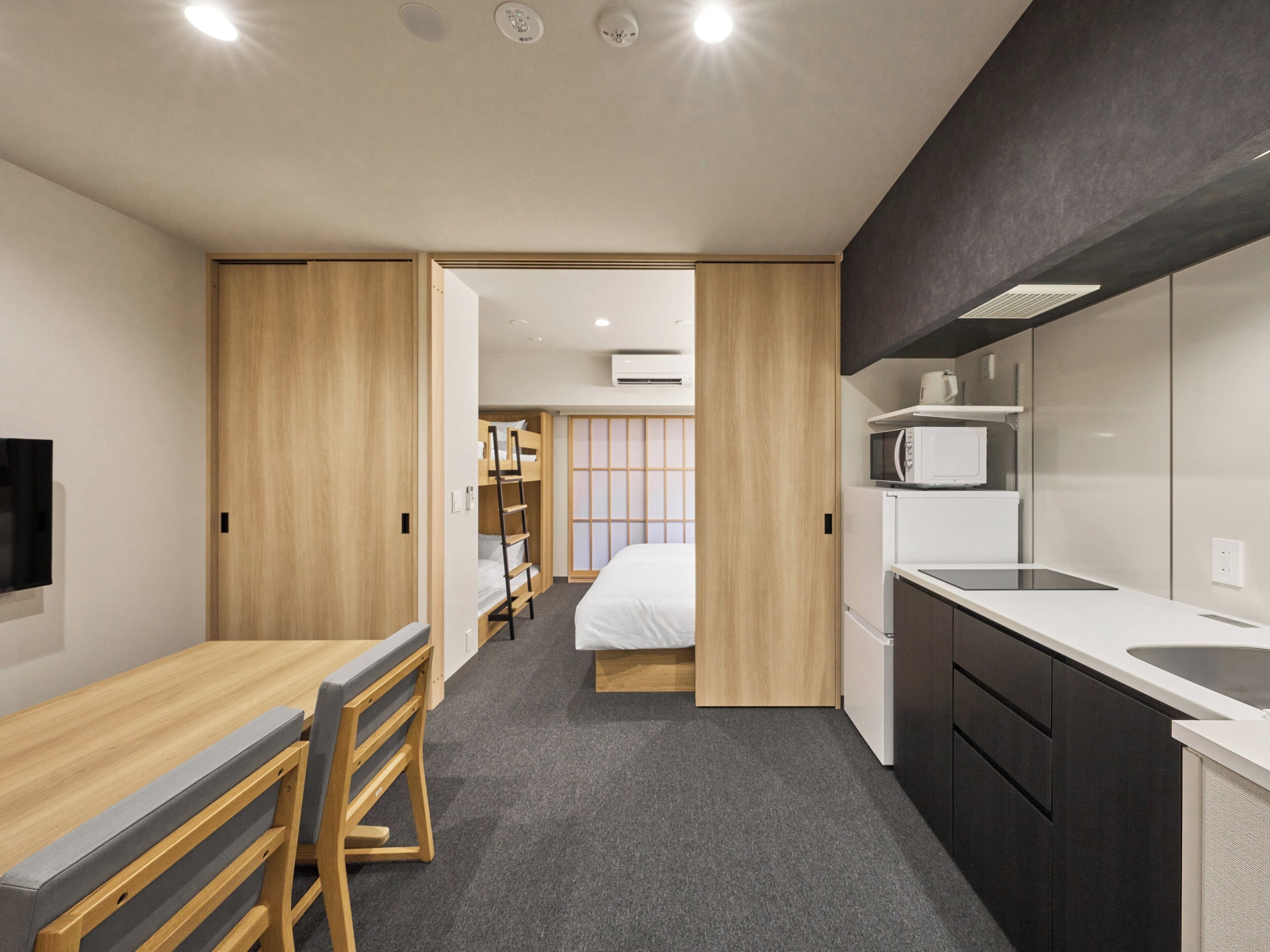A compact hotel room with a kitchenette, a bed with a futon, a wooden desk, and a TV.