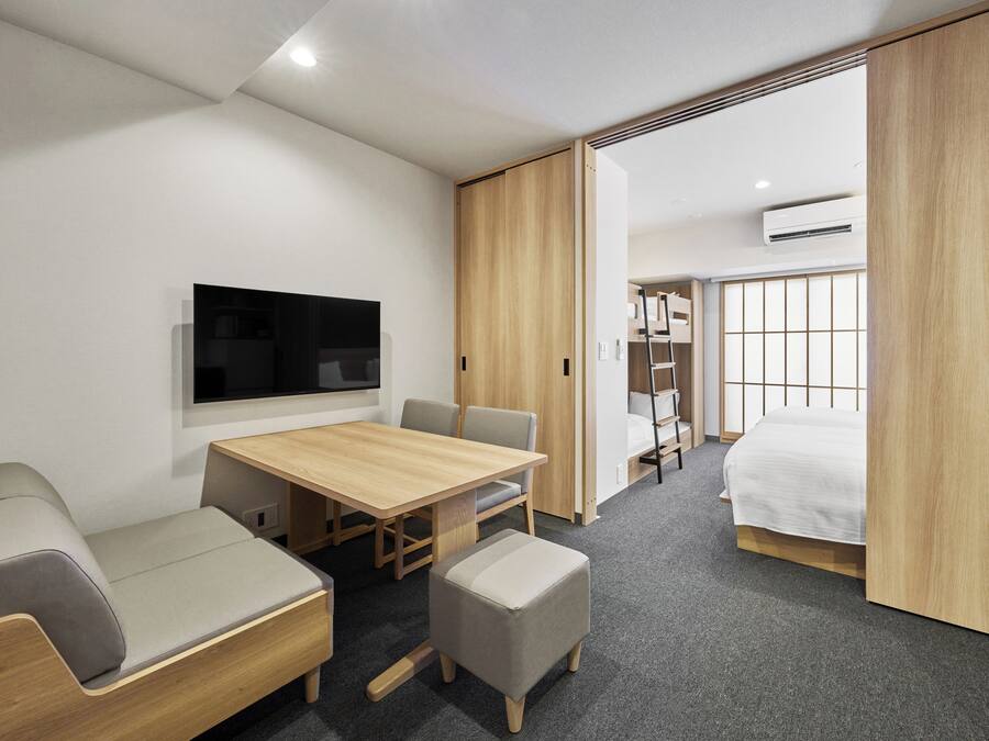 A modern hotel room with a wooden dining table, a sofa, a small ottoman, a TV mounted on the wall, a bunk bed with a ladder, and an air conditioning unit.