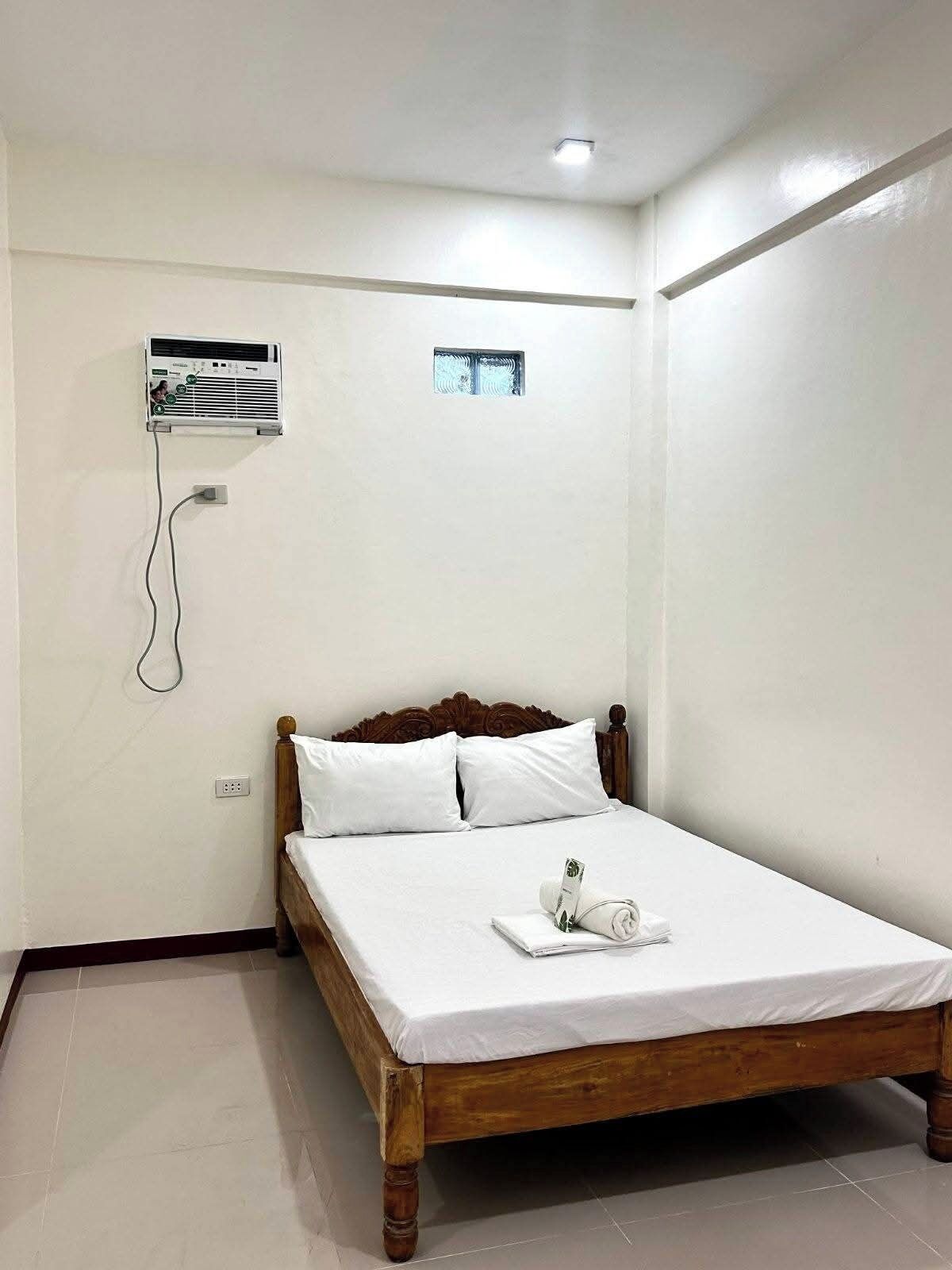 Een enkel bed met houten frame en witte beddengoed, een wandairconditioner en een klein raam.