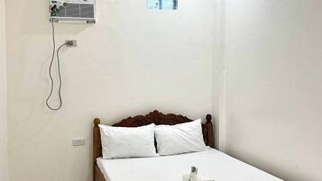Een enkel bed met houten frame en witte beddengoed, een wandairconditioner en een klein raam.