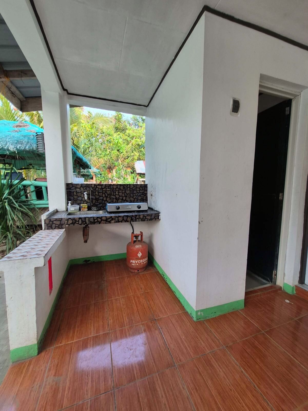 A small kitchen area with a gas stove, a gas cylinder, and a window overlooking greenery.