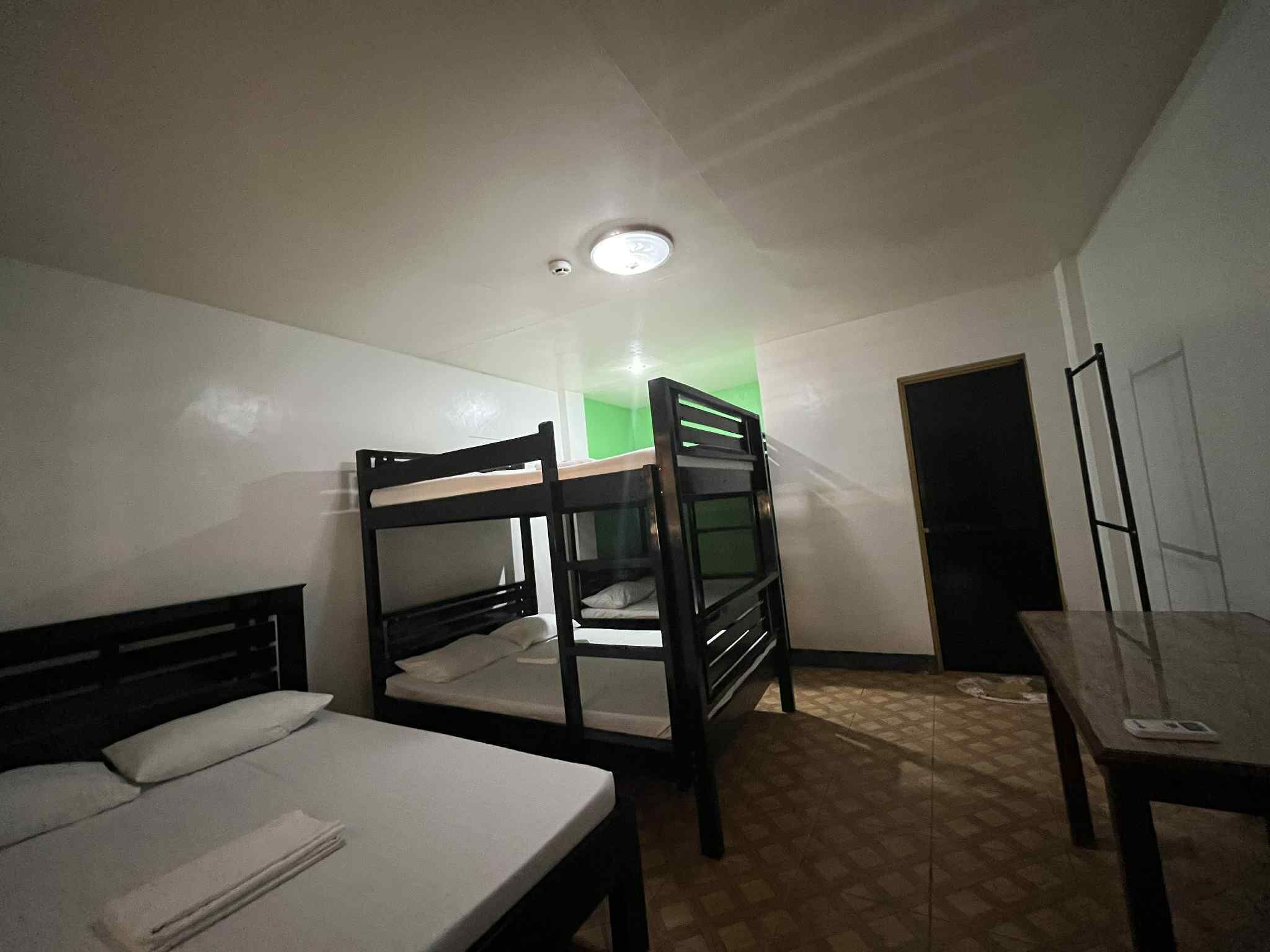 Une petite pièce faiblement éclairée, avec des lits superposés, un lit simple, une table et une porte.
