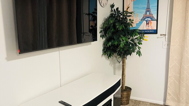 A living room with a flat-screen TV mounted on the wall, a white entertainment unit below, a potted plant, a clock, and a poster of the Eiffel Tower. - Spacious2 - bedroom condo in tranquil (Reynoldsburg)