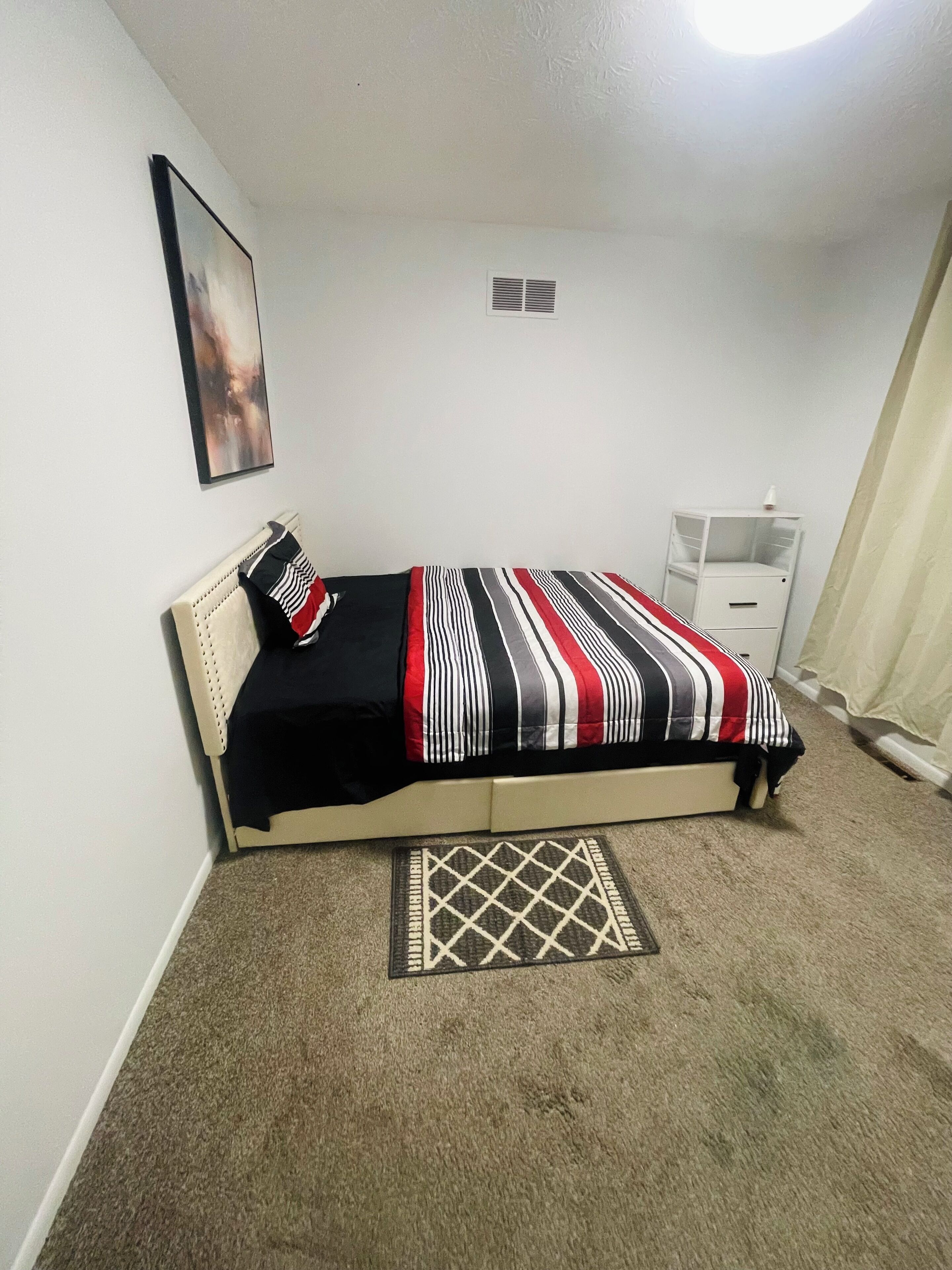 Ein Schlafzimmer mit einem Bett, einem Nachttisch, einer kleinen Kommode, einem Teppich und einem Bild an der Wand.
