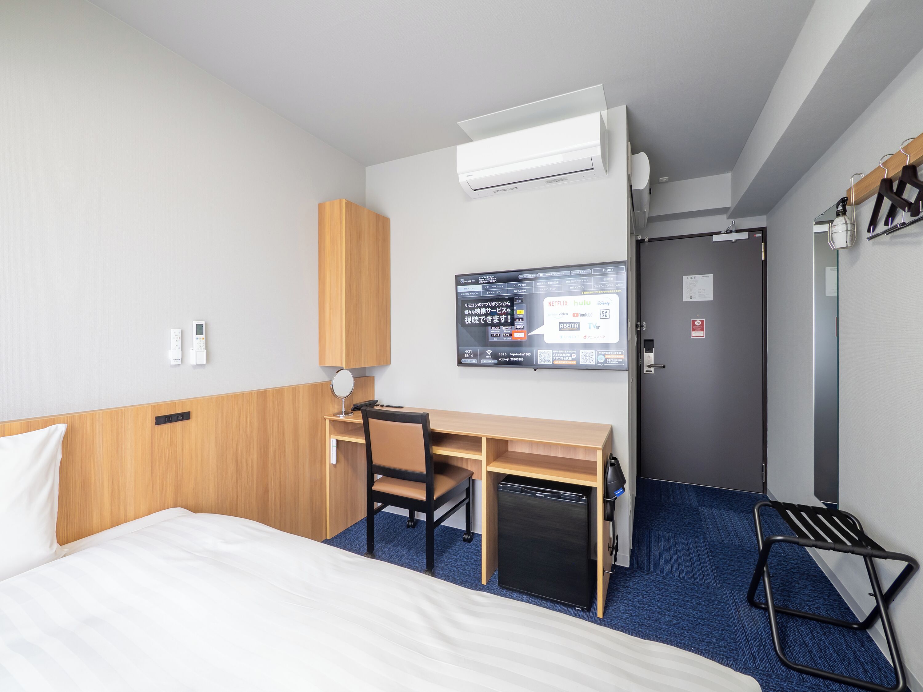 A hotel room with a bed, a desk with a chair, a small refrigerator, an air conditioning unit, a wall-mounted TV, and a door with a handle.