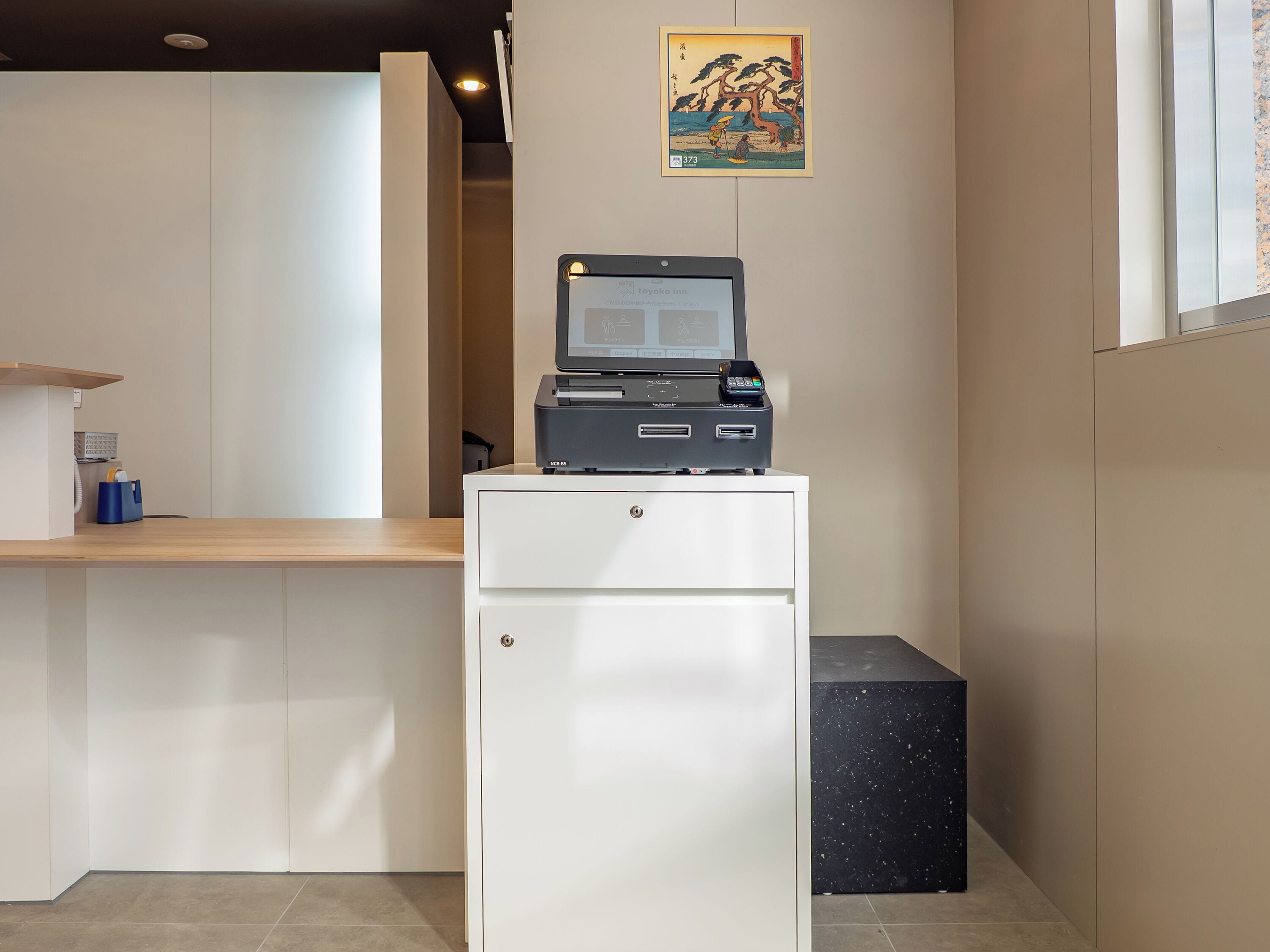Un mostrador de recepción con una pantalla digital, un refrigerador pequeño y un cuadro colgado en la pared.