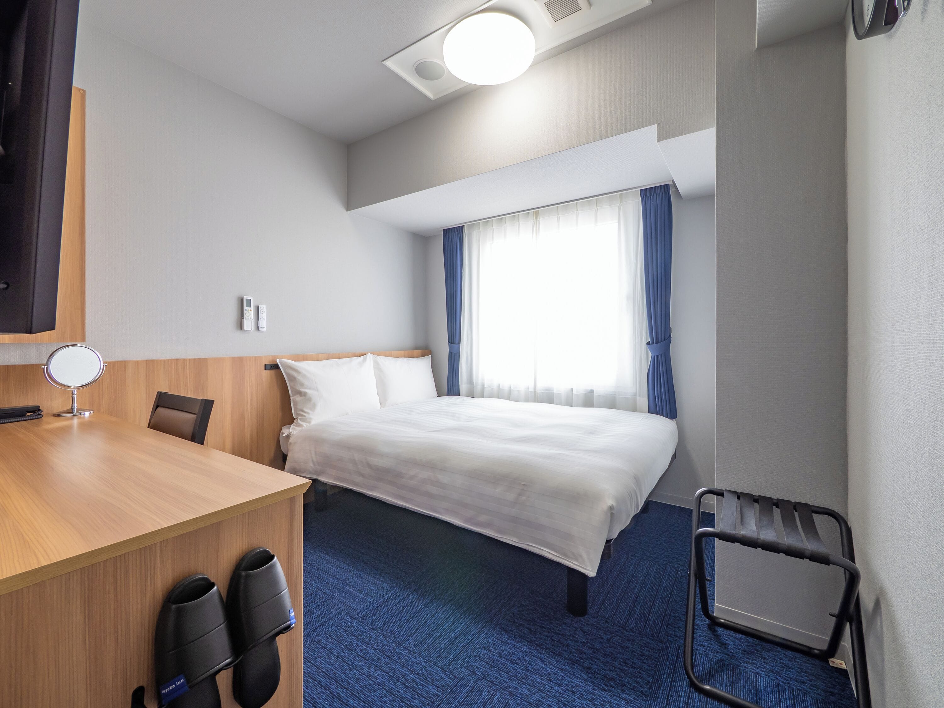 A hotel room with a bed, a desk with a mirror, a chair, a blue carpet, and a window with curtains.