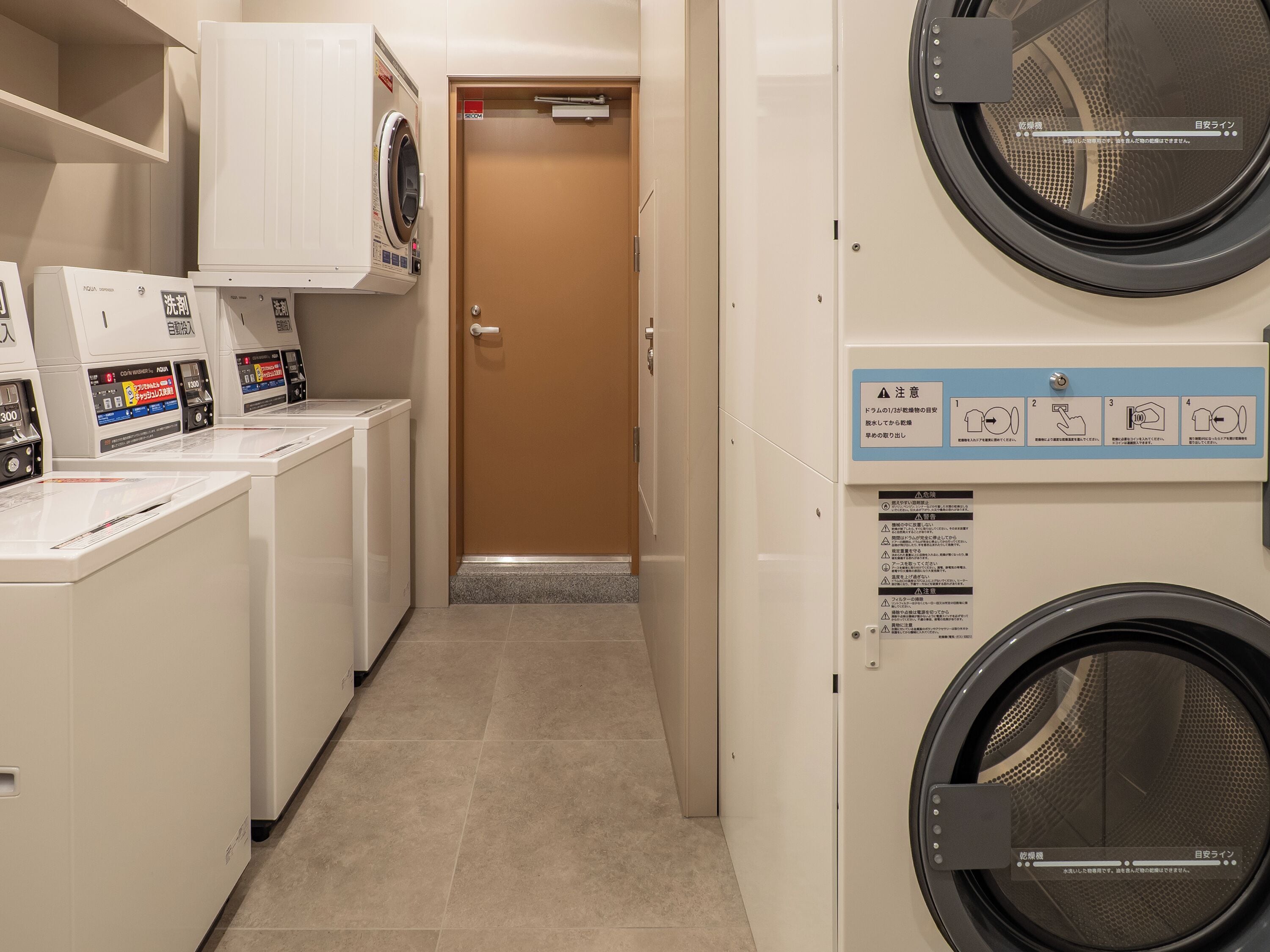 A laundry facility with multiple washing machines and dryers, a door with a sign, and a tiled floor.