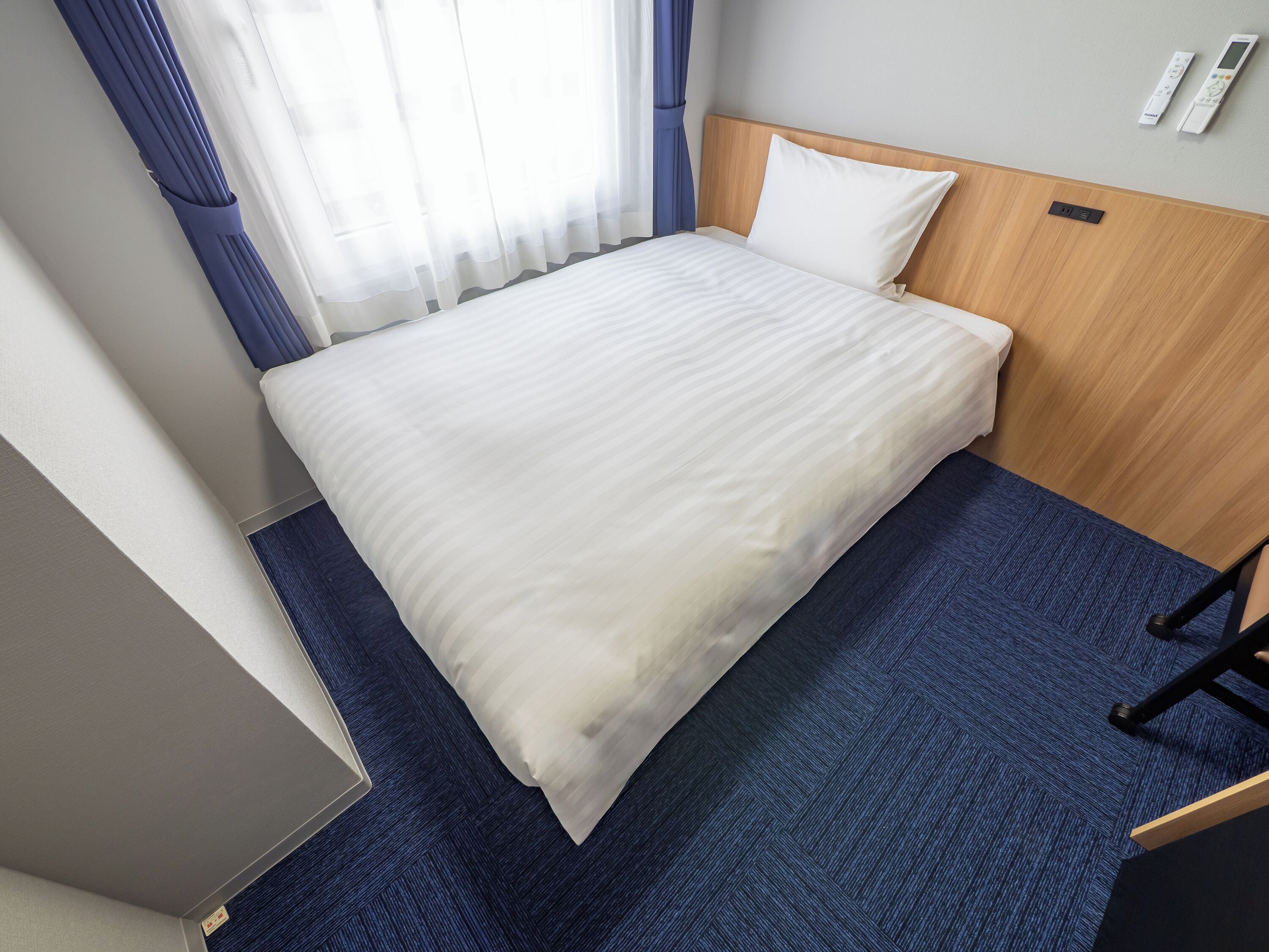 A hotel room with a bed, a wooden headboard, a blue carpet, and a window with curtains.