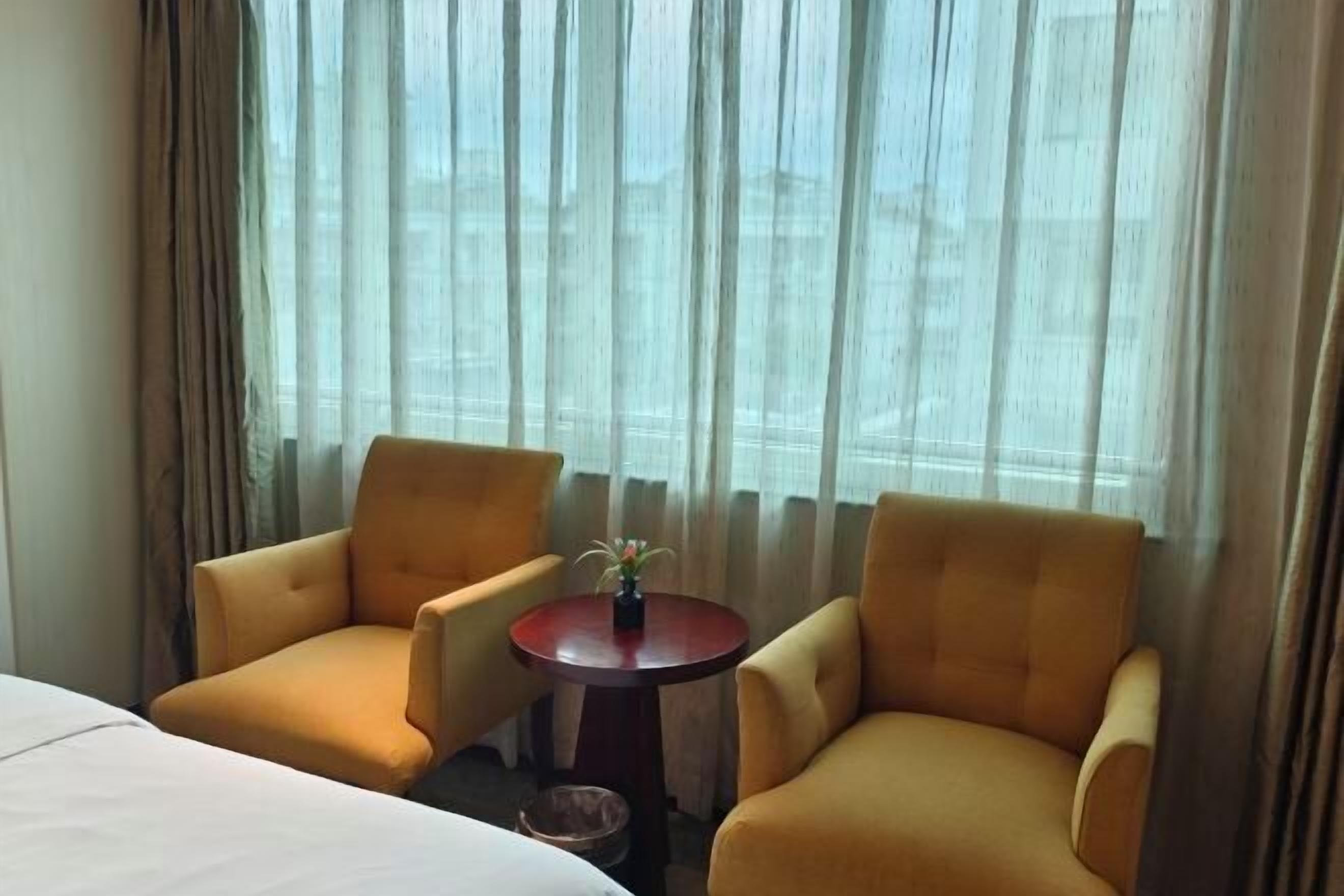 Habitación de hotel con cama, dos sillones amarillos, una mesita redonda con una planta y una ventana con cortinas translúcidas.