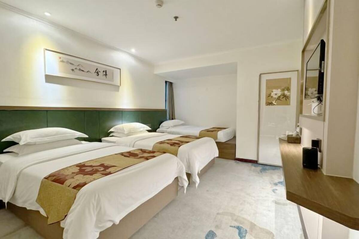 A hotel room with three beds, a green headboard, a flat-screen TV, and a framed artwork on the wall.