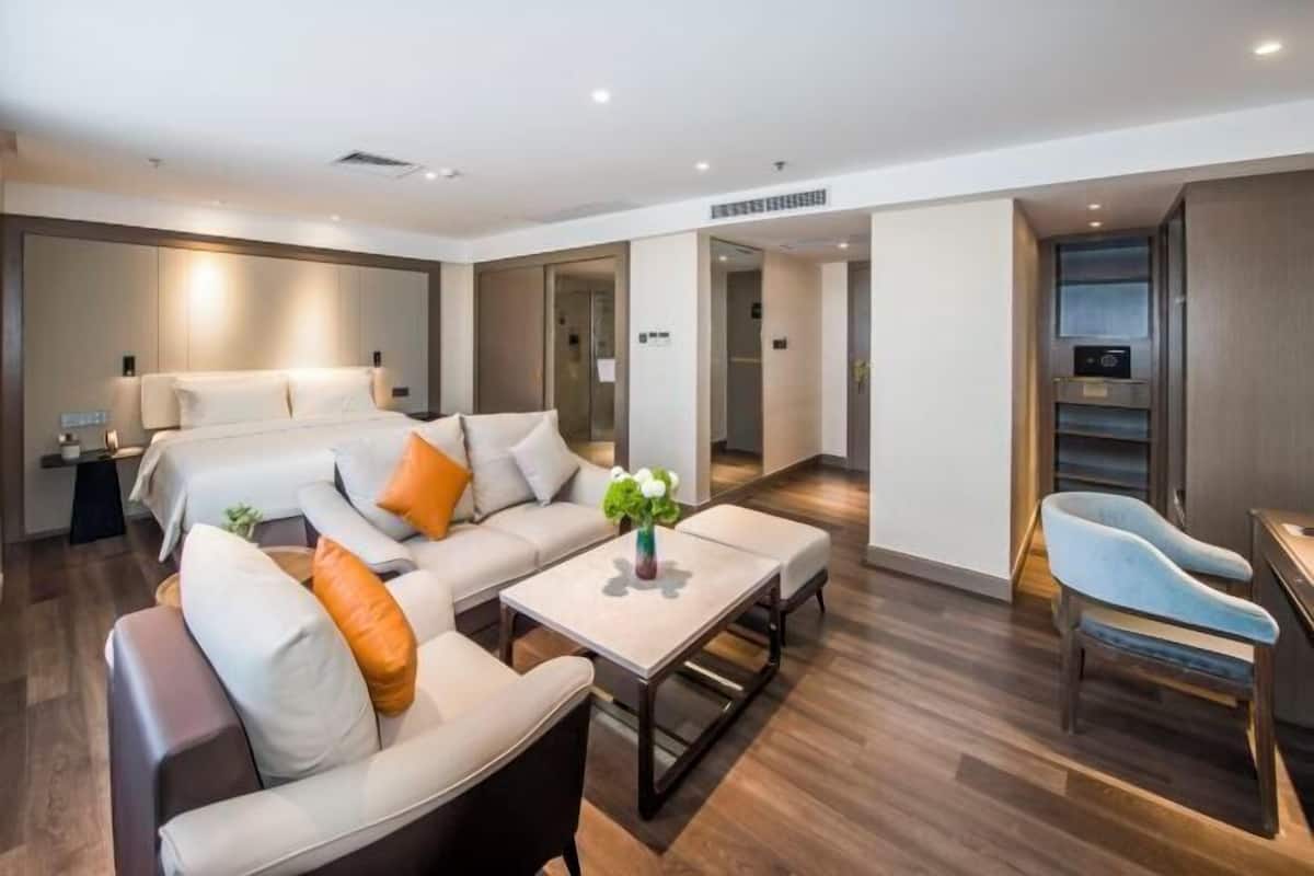 Een moderne hotelkamer met een bank, een salontafel, een bureau en een stoel. Achtergrond is een bed en een deur die leidt naar een andere kamer.
