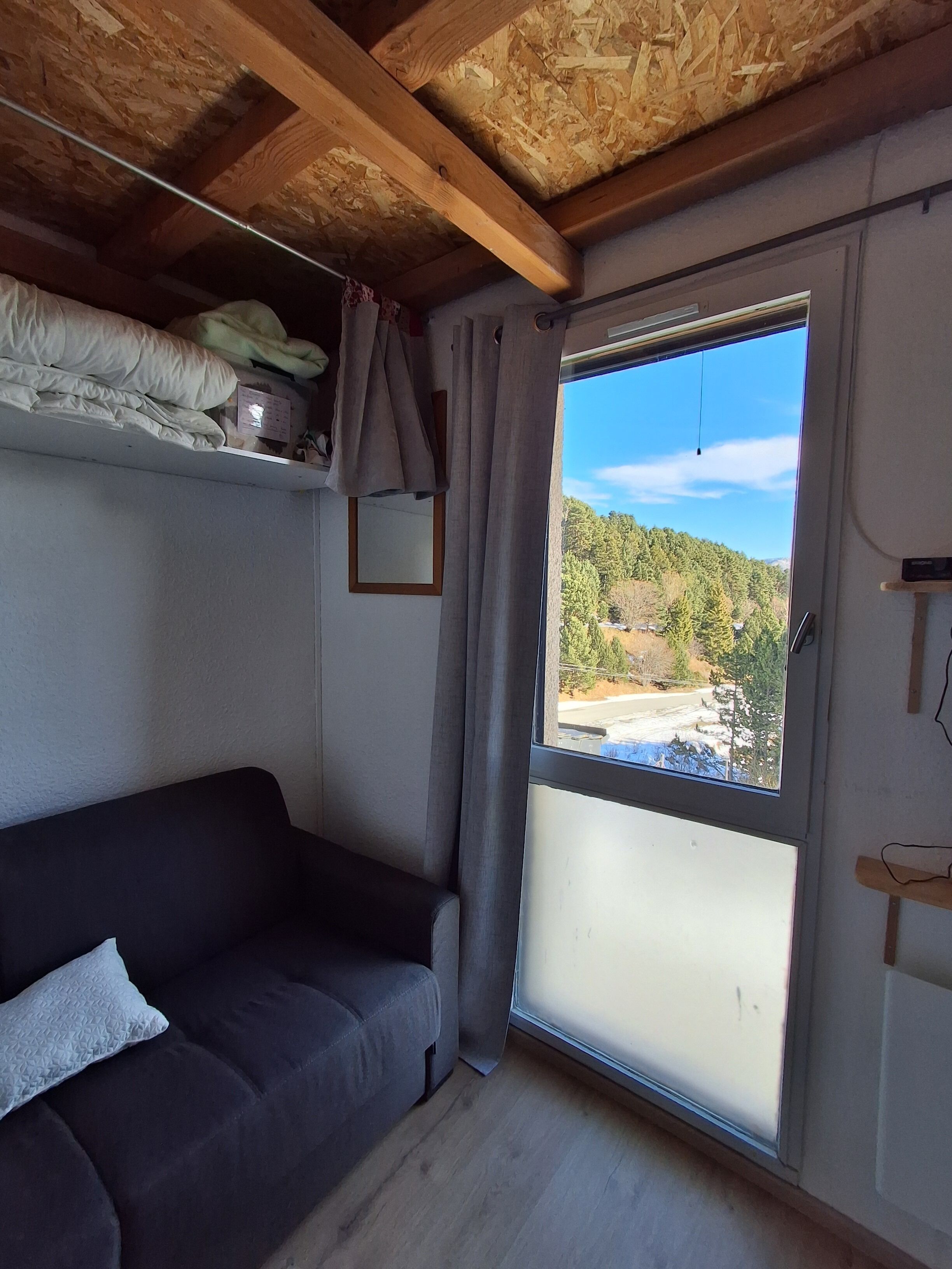 Una habitación con un sofá de color oscuro, techo de madera, ventana con vistas a un área boscosa y una puerta con marco de madera.